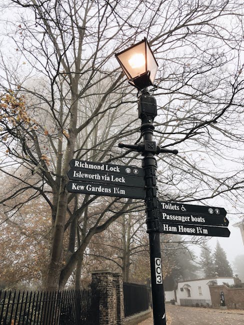 A black Victorian-style lamppost with a lit lantern at the top is situated in a garden or park area with partially leafless trees in the background, indicating autumn or early winter. Attached to the 